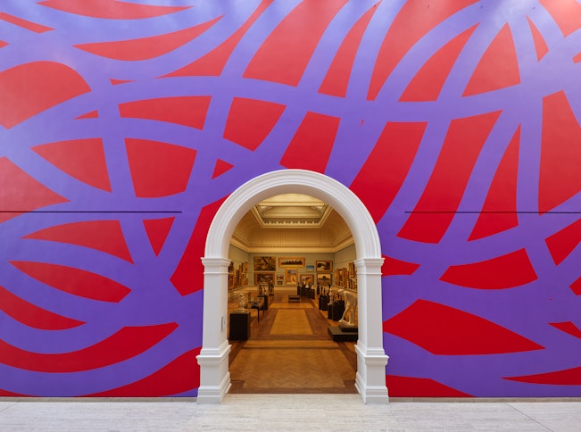 A large wall with purple looping lines on a red background. An arched doorway leads into a gallery filled with paintings and sculptures.