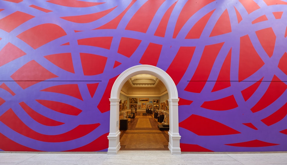 A large wall with purple looping lines on a red background. An arched doorway leads into a gallery filled with paintings and sculptures.