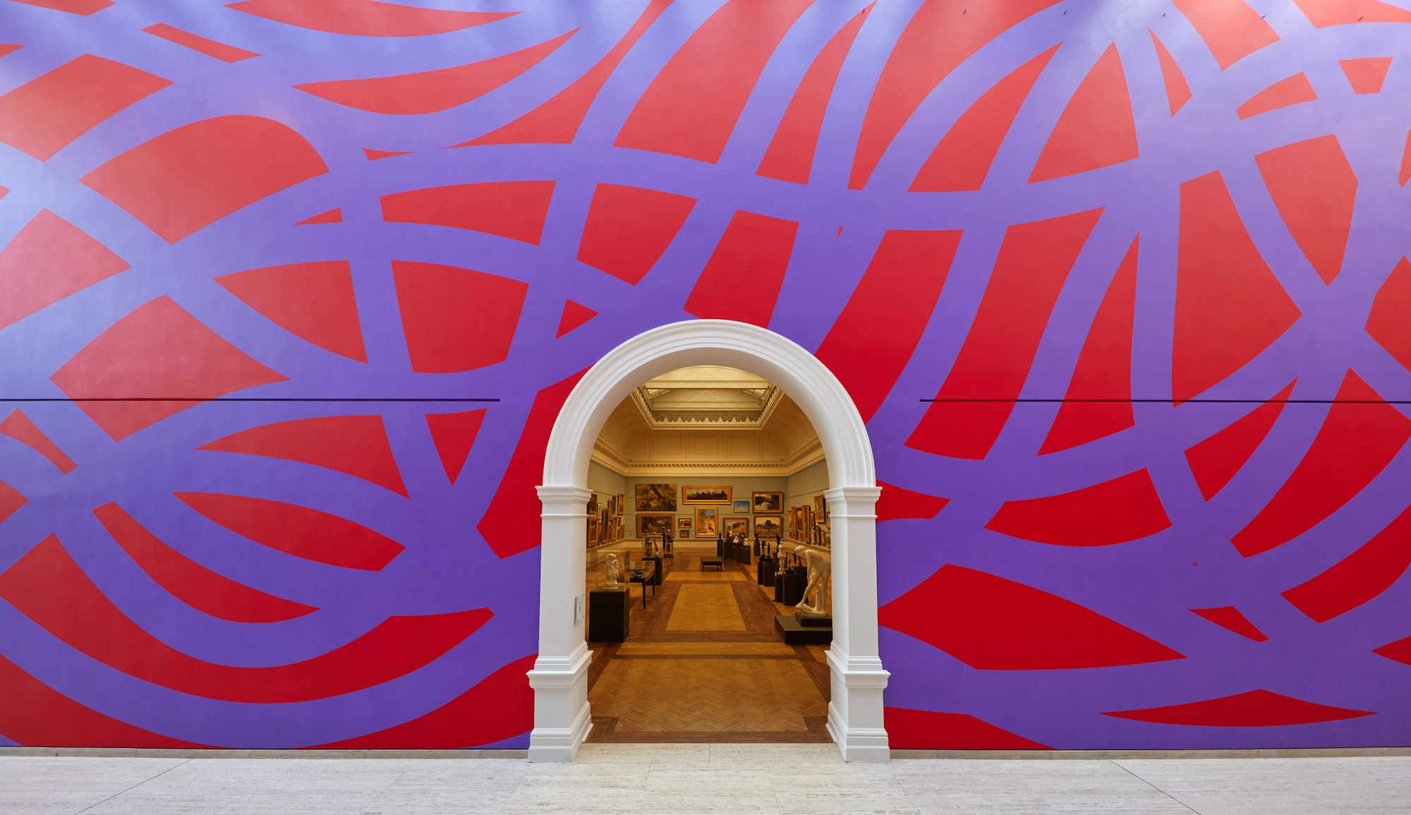 A large wall with purple looping lines on a red background. An arched doorway leads into a gallery filled with paintings and sculptures.