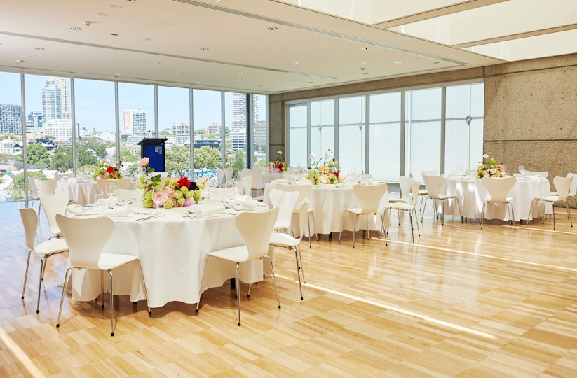 A room with a large window looking over a city. The room has a lecturn and several round cloth-covered and decorated tables with chairs