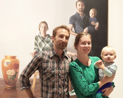 Two people, one holding a baby, stand in front of a painting in which all three are depicted