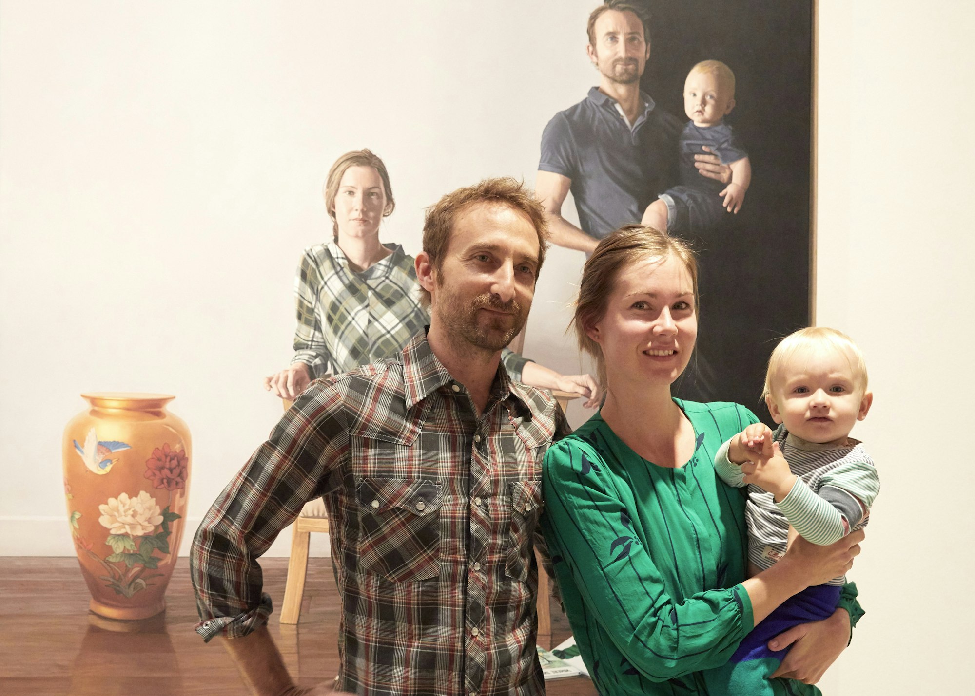 Two people, one holding a baby, stand in front of a painting in which all three are depicted