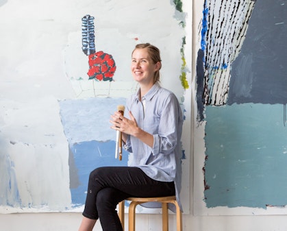 A person holding paintbrushes sits on a stool  in front of paintings on a wall