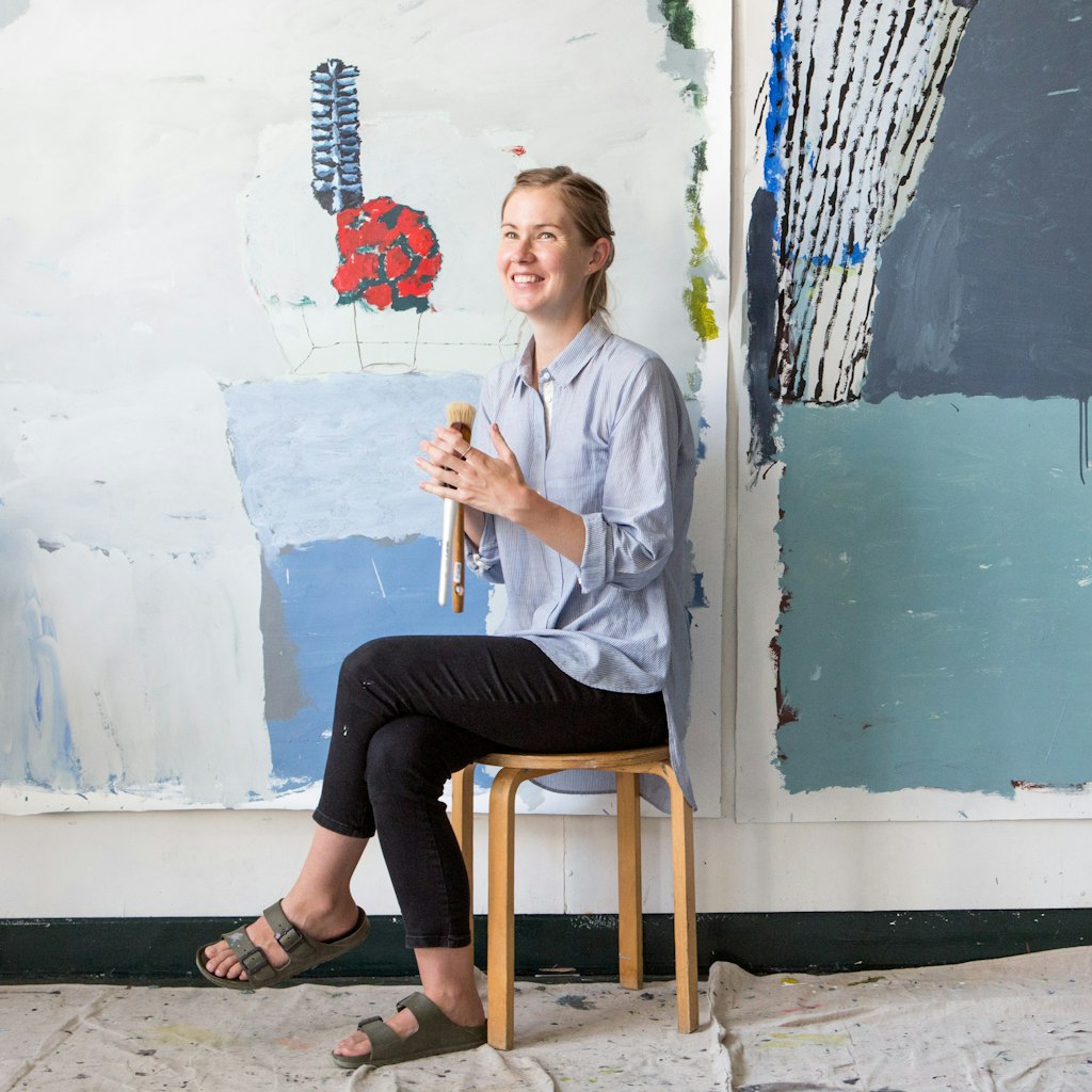 A person holding paintbrushes sits on a stool in front of paintings on a wall