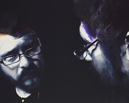 A bearded person wearing glasses looks at their own reflection