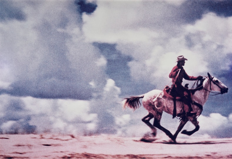 A person rides a horse across bare gound with large white clouds behind