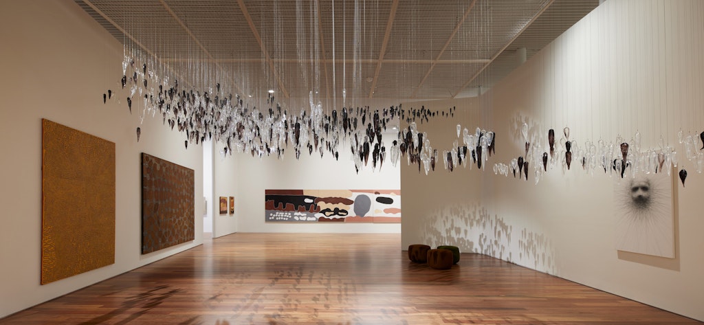 A gallery space with paintings and drawings on the walls and a large glass sculpture hanging from the ceiling