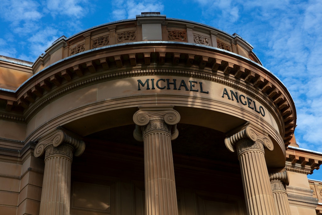 The top of a sandstone building with columns, above which is written 'Michael Angelo'