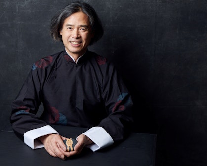 A person with dark, chin-length bobbed hair, dressed in a black high-collared robe with wide white cuffs, sits holding a small wrapped object