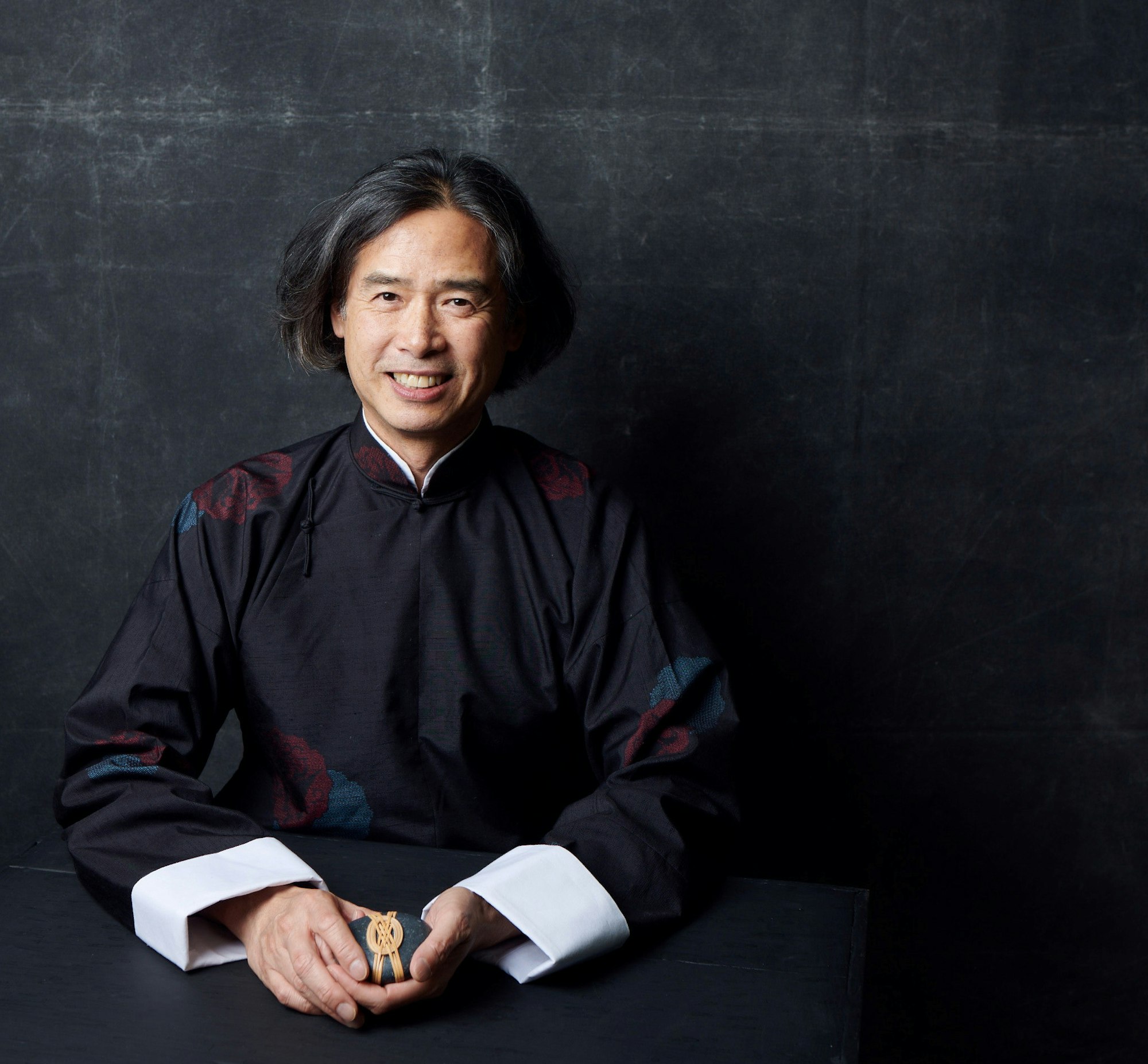 A person with dark, chin-length bobbed hair, dressed in a black high-collared robe with wide white cuffs, sits holding a small wrapped object
