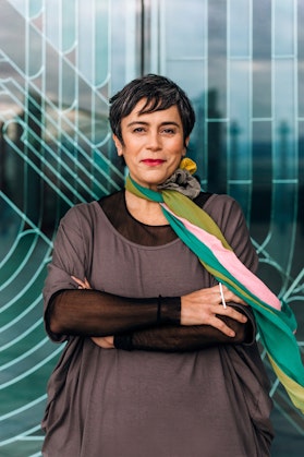 A person with short dark hair wearing a colourful long scarf around their neck and black-and-grey layered clothes, stands with arms crossed
