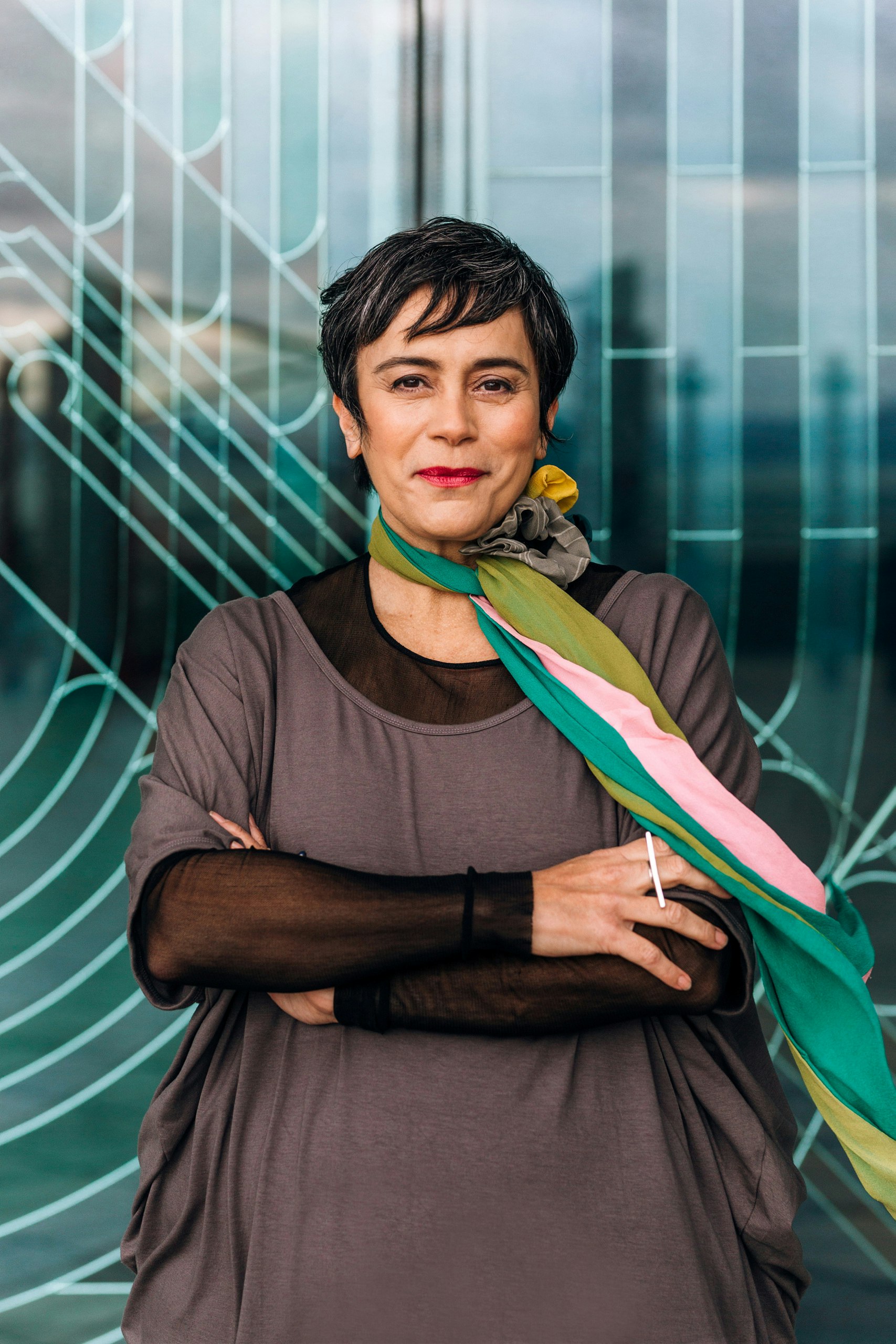 A person with short dark hair wearing a colourful long scarf around their neck and black-and-grey layered clothes, stands with arms crossed