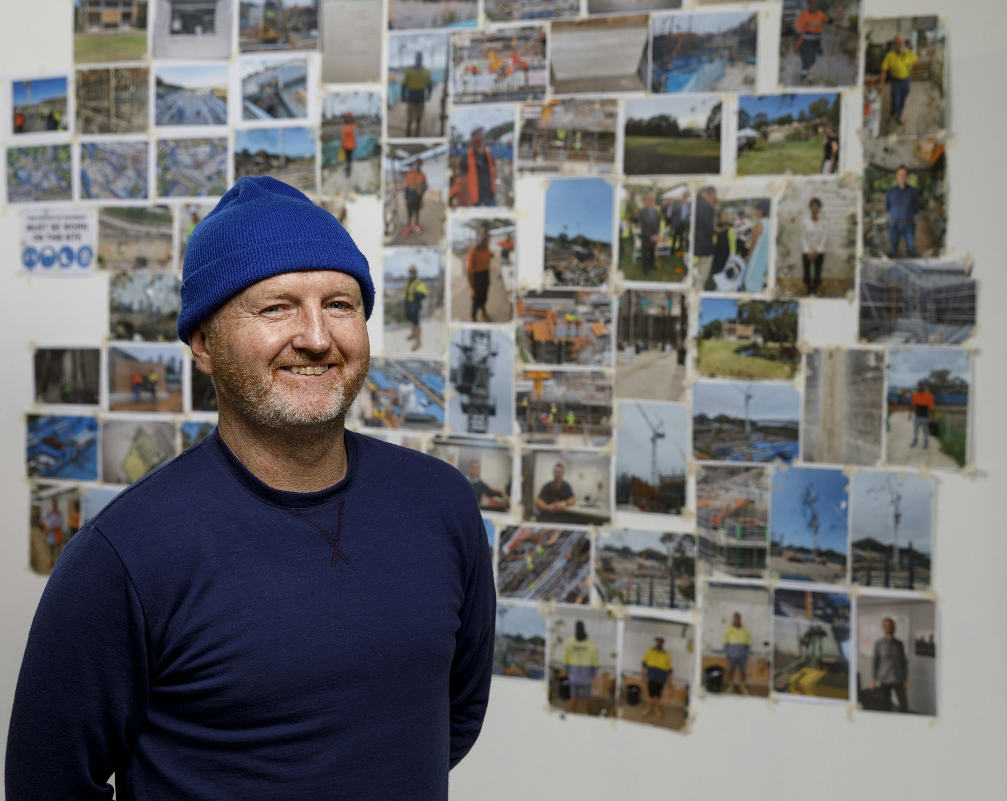 A person wearing a blue beanie stands in front of a wall holding a multitude of unframed photos.
