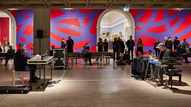 Two people sit in front of musical equipment, opposite each other, while people watch on in a gallery with a red and purple painted wall.