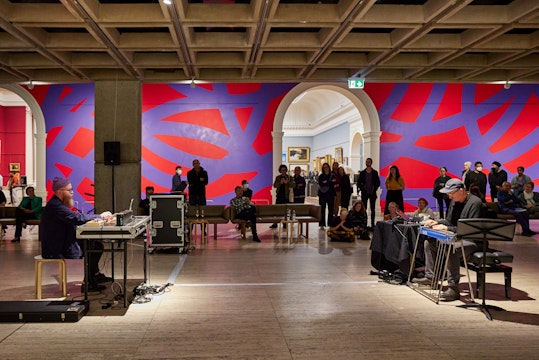 Two people sit in front of musical equipment, opposite each other, while people watch on in a gallery with a red and purple painted wall.