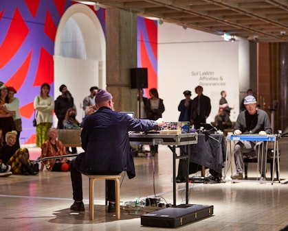 Two people sit in front of musical equipment, opposite each other, while people watch on in a gallery with a red and purple painted wall.