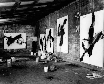 A black and white photograph of a warehouse studio with five large paintings hung on the walls. A person stands in the far corner.