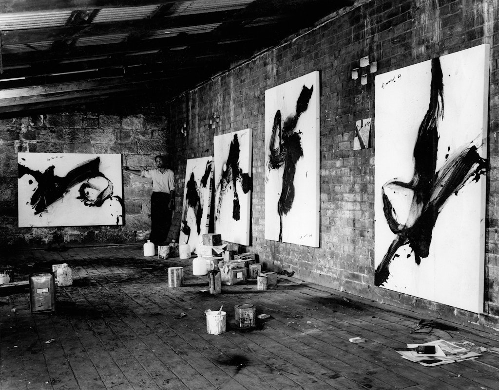 A black and white photograph of a warehouse studio with five large paintings hung on the walls. A person stands in the far corner.