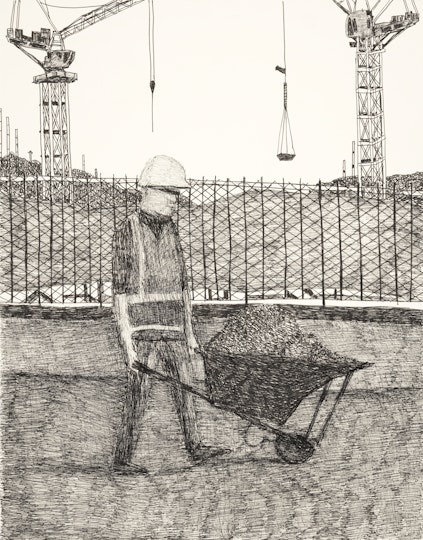 A person in a hard hat pushes a wheelbarrow in front of a fence and two giant cranes