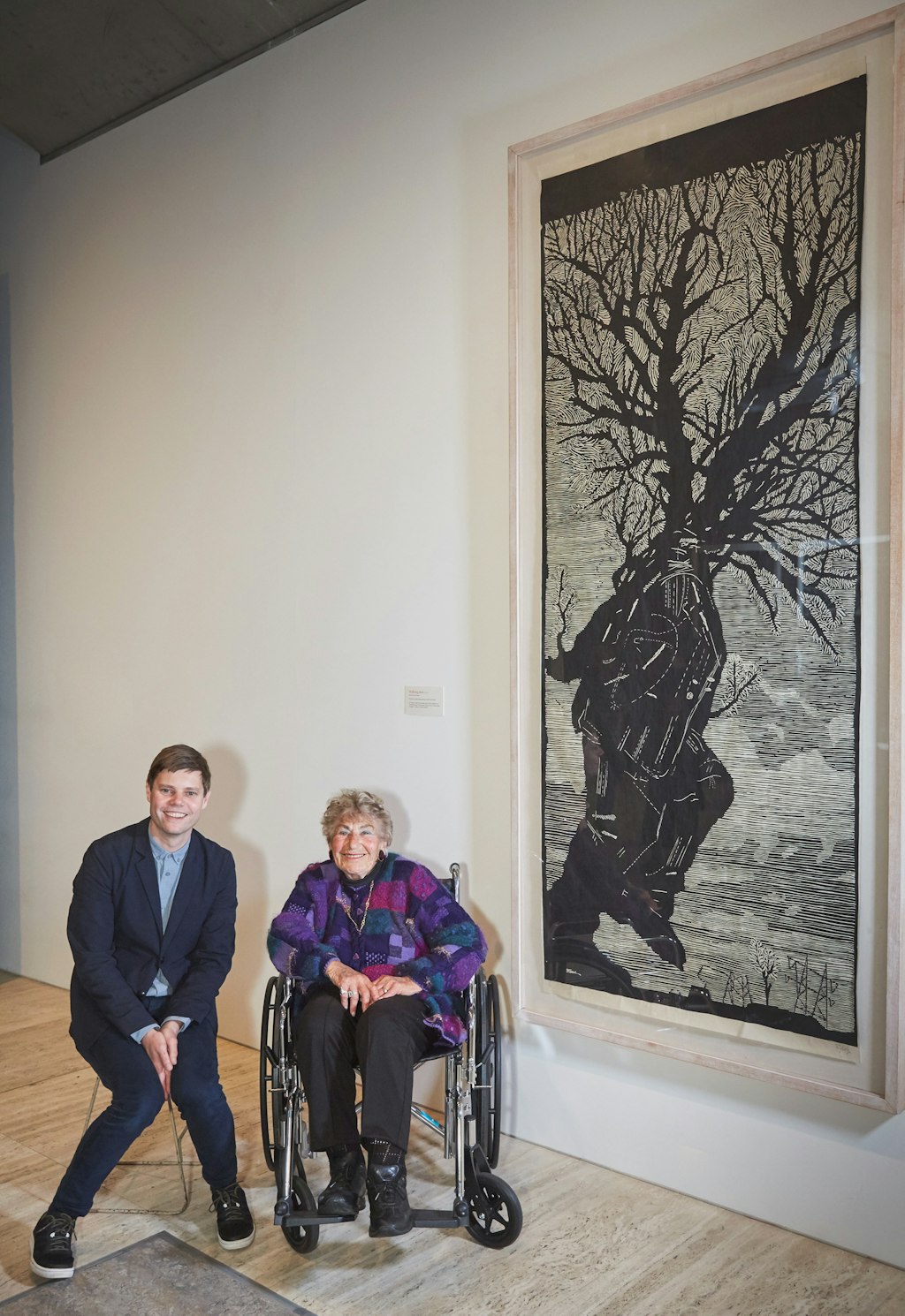 Two people sit next to a large black-and-white framed artwork depicting a figure that is part human, part tree