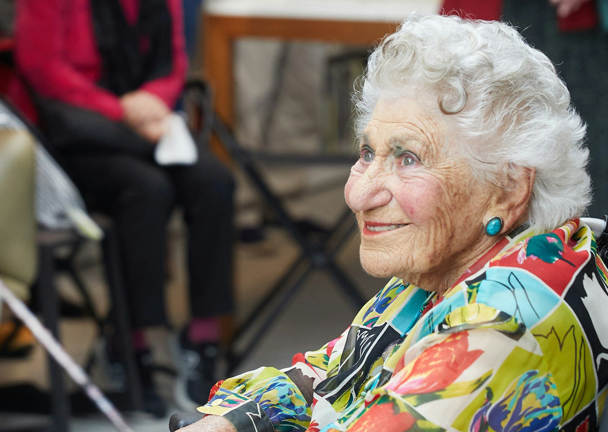 A smiling person with short wavy white-grey hair, blue earrings and colourful clothing