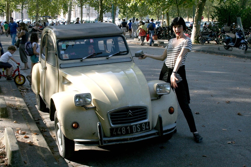 A person stands next to a vintage car on a tree-lined street