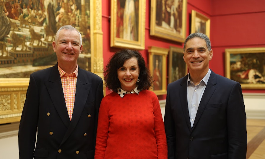 Three people stand in a red-walled space hung with gold-framed paintings