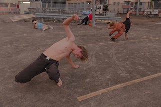A carpark rooftop. Grey concrete, covered in tyre marks, and the yellow outlines of parking spaces. In the middle of the space, a diverse group of four performers are mid dynamic movement. Backs arch, arms lift, one person leans against a lamp post, two others stretch out on the concrete