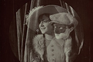 A person in a wide-brimmed feathered hat and other wearing a peaked cap with their upper face covered by a mask