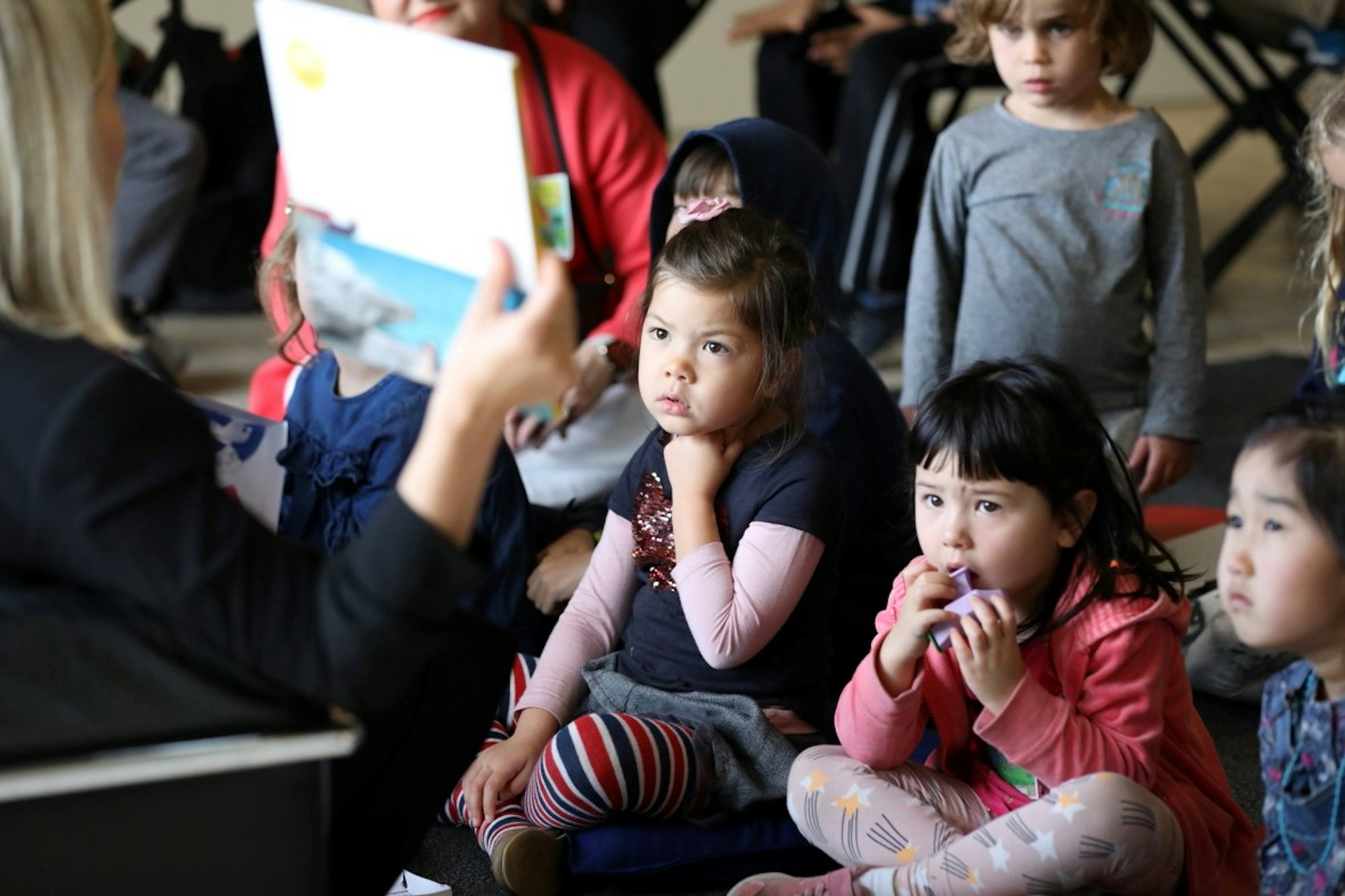 Children sit on the floor looking up at a person who is holding a book
