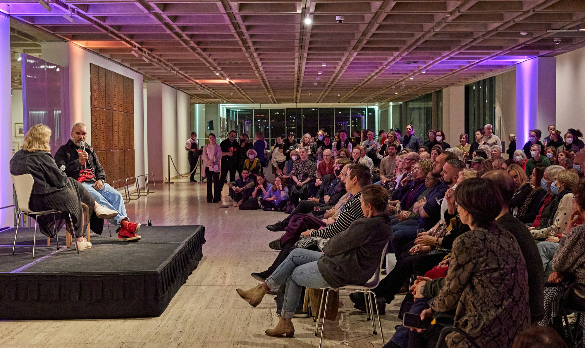 Two people seated on a low stage in a large room in front of an audience
