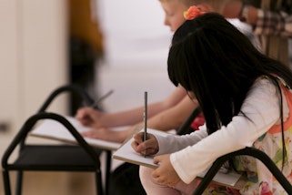 Two seated children drawing with pencils on paper on a clipboard