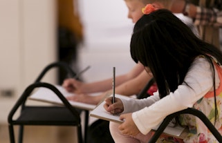 Two seated children drawing with pencils on paper on a clipboard