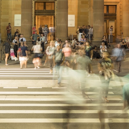 People crossing a pedestrian crossing in front of a building where other groups of people wait and move around