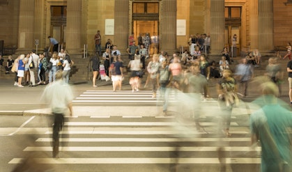 People crossing a pedestrian crossing in front of a building where other groups of people wait and move around