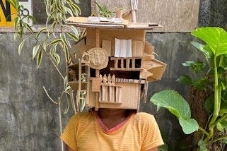 A person with a house-like object made from cardboard on their head like a mask
