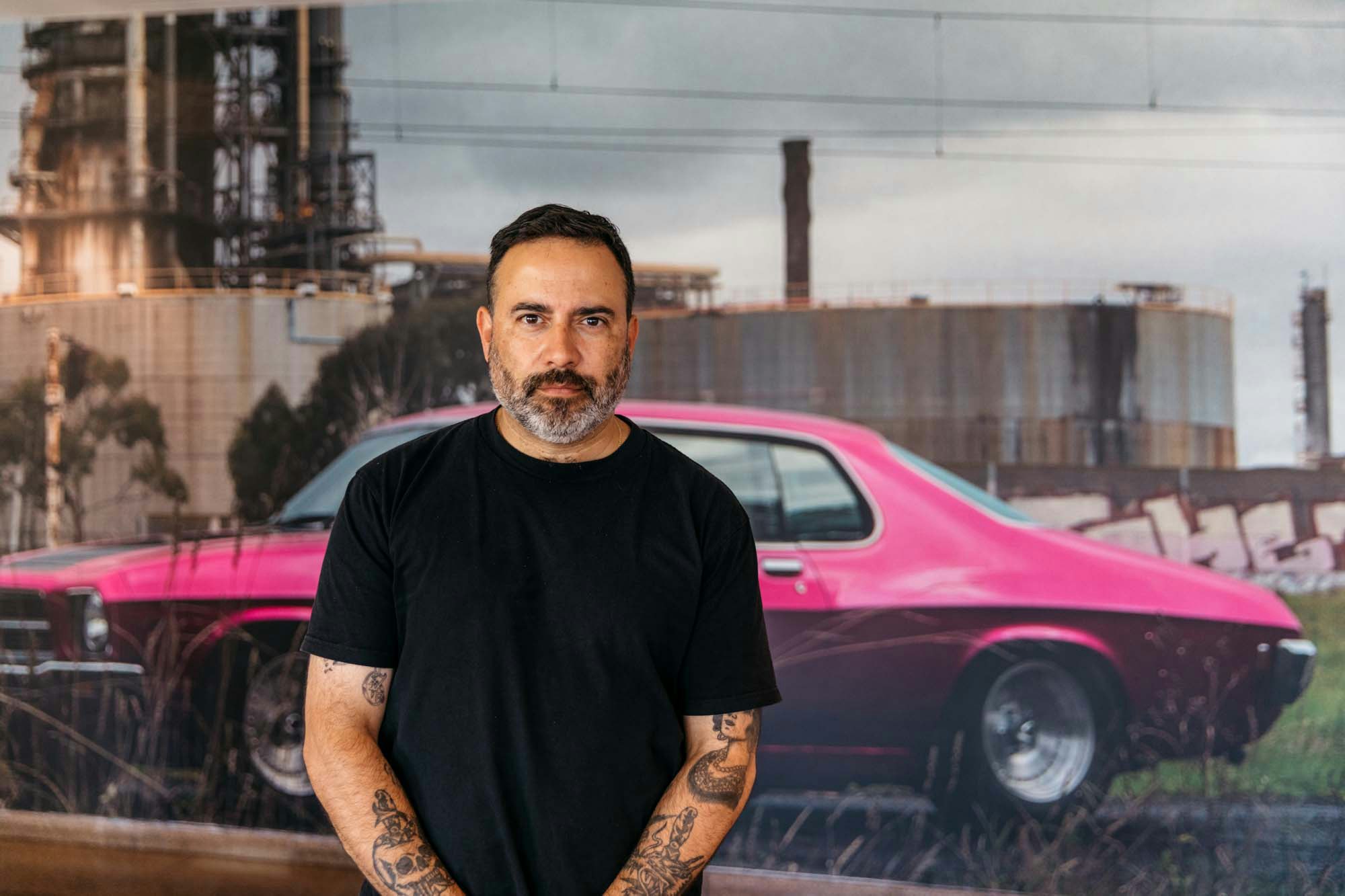 A bearded person stands in front of a photo of a pink car in an industrial landscape