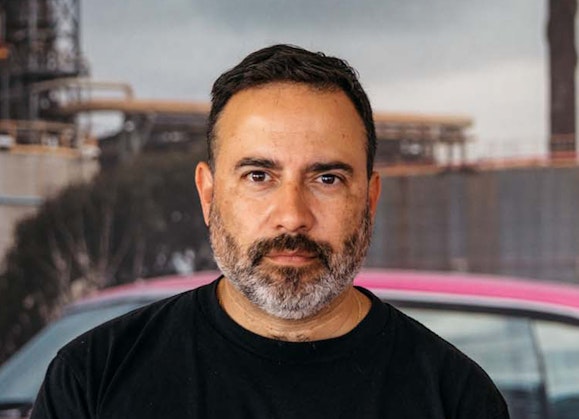 A bearded person stands in front of a photo of a pink car in an industrial landscape
