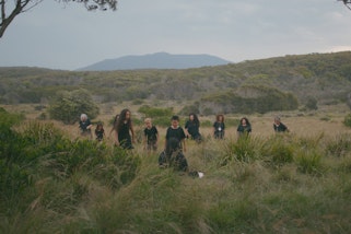 A group of 10 people dressed in black moving through a landscape of grass and trees