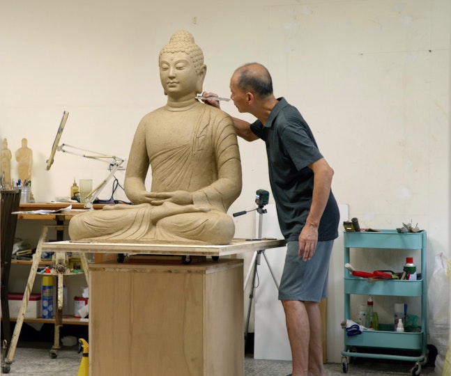A person uses a small tool to work on a large seated Buddha sculpture