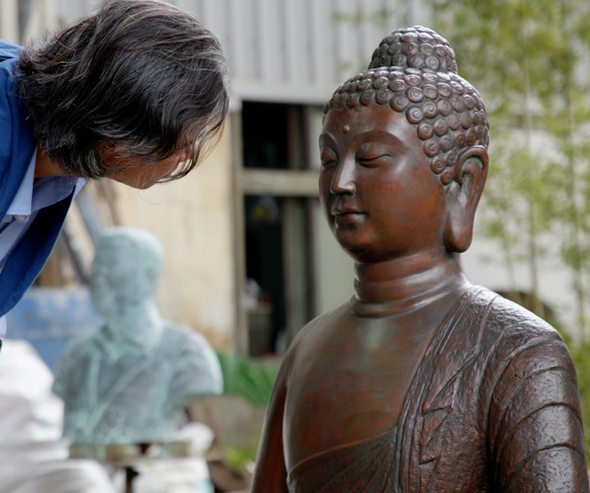 A person leans over to inspect a bronze Buddha sculpture