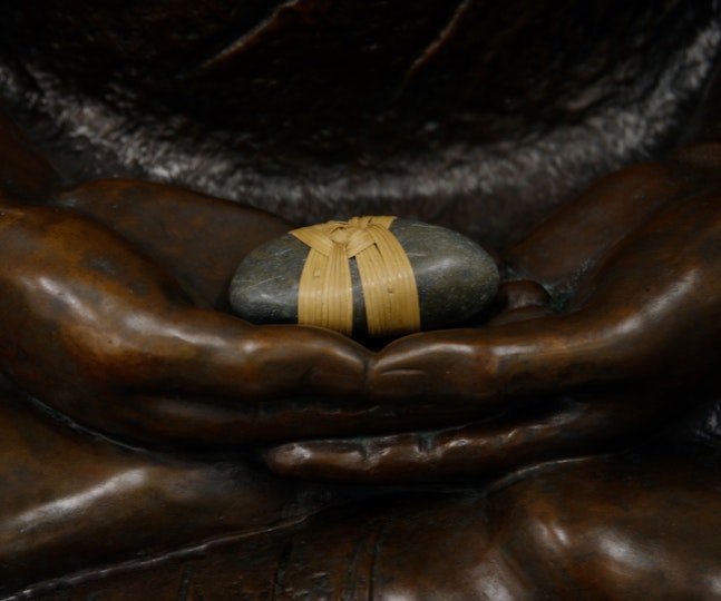 A small grey stone, with two yellowish bands wrapped and knotted around it, sits within the upturned palms of a bronze sculpture