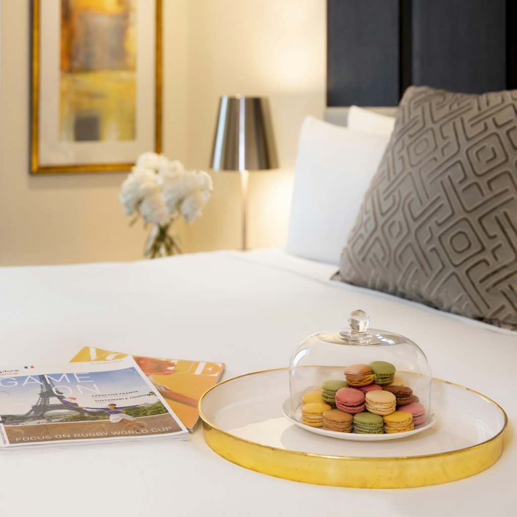 A plate of macarons and two magazines on a bed. On the bedside table is a lamp and vase of flowers.