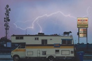 A campervan, with a dog on the roof, is parked between a telecommunications tower and a neon sign. Lightning cracks across a purplish sky.