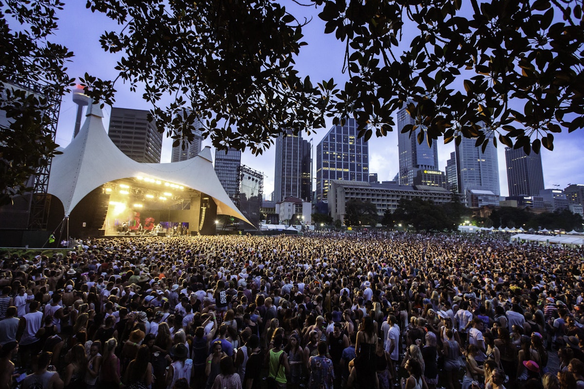 Crowds at dusk in front of a covered stage with tall city buildings behind