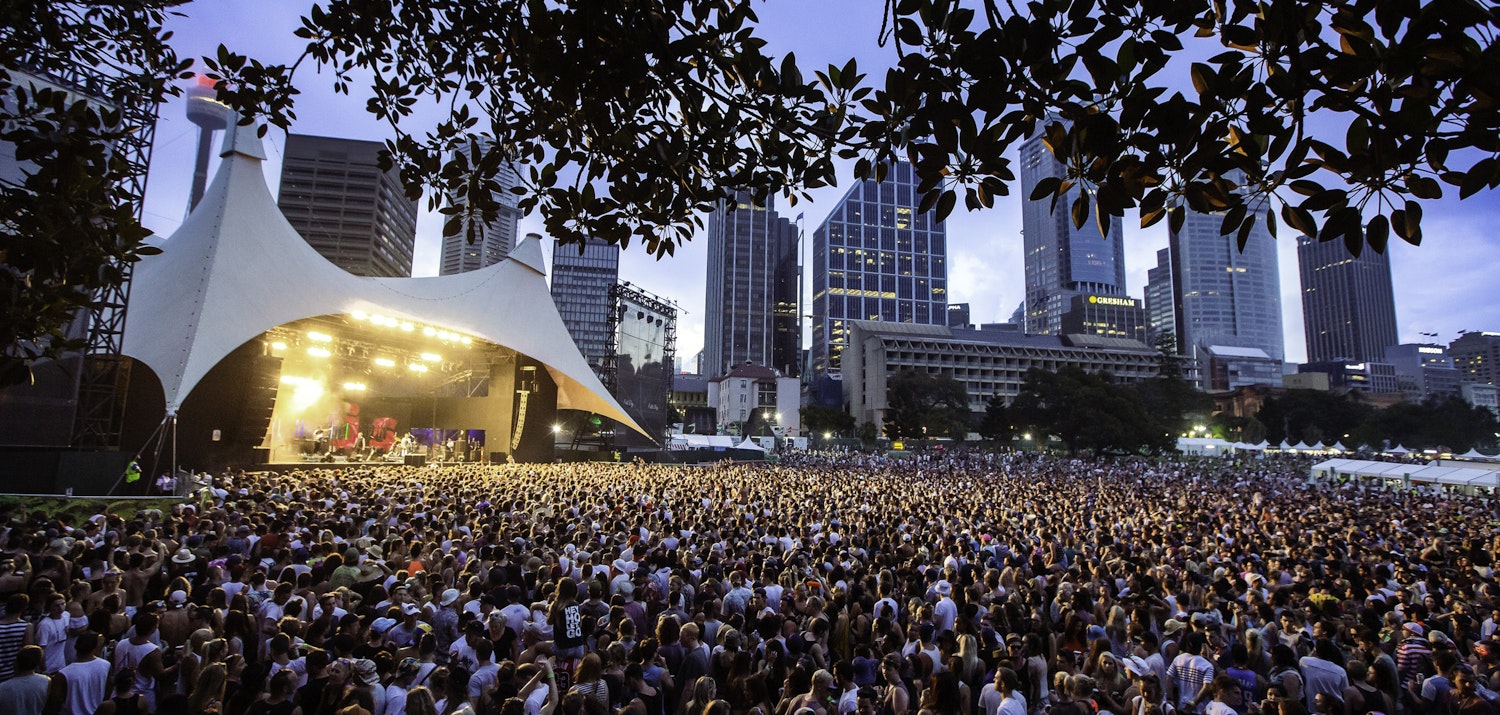 Crowds at dusk in front of a covered stage with tall city buildings behind