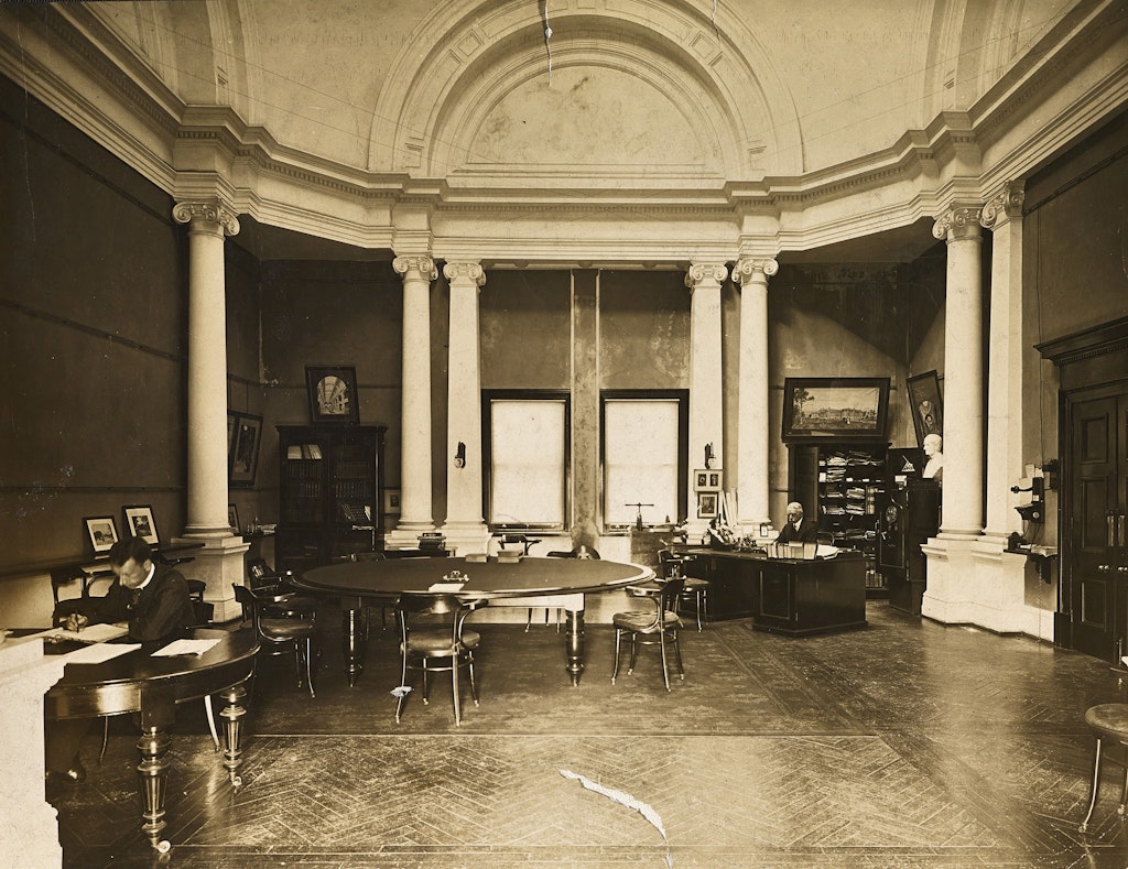 A room with columns and ornate upper walls, in which there are a few tables, a desk and some small cabinets