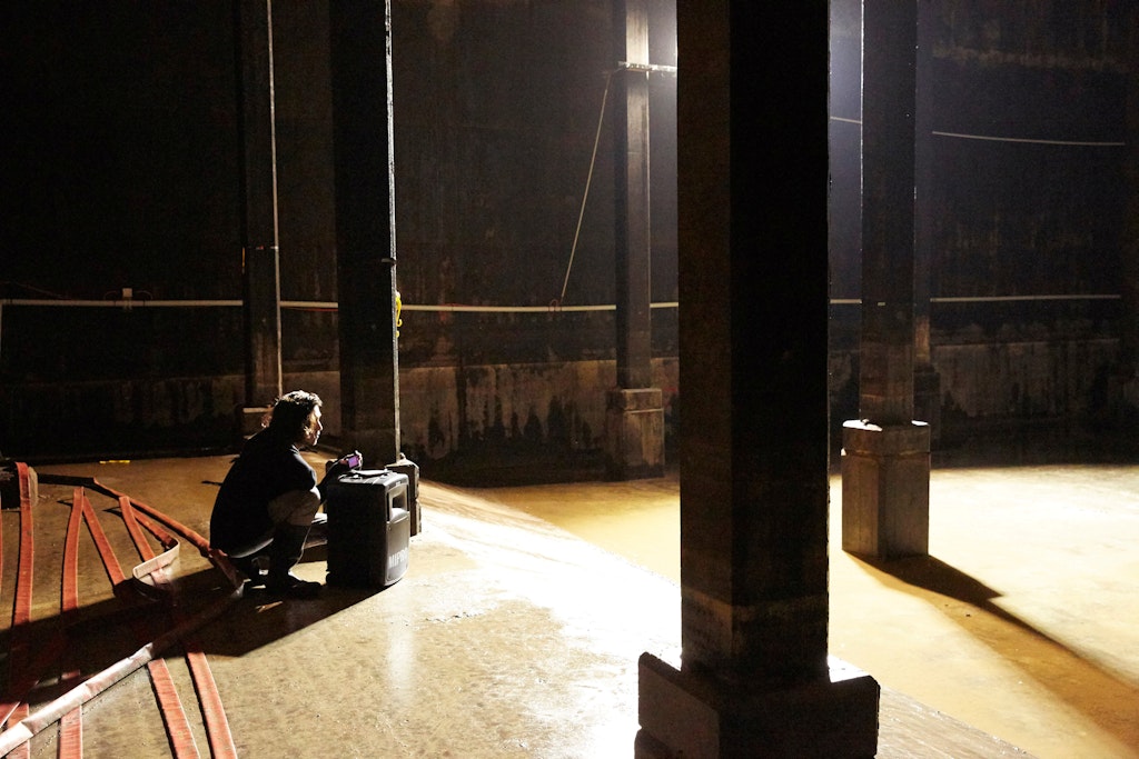 A person squats next to an audio speaker within a room with columns