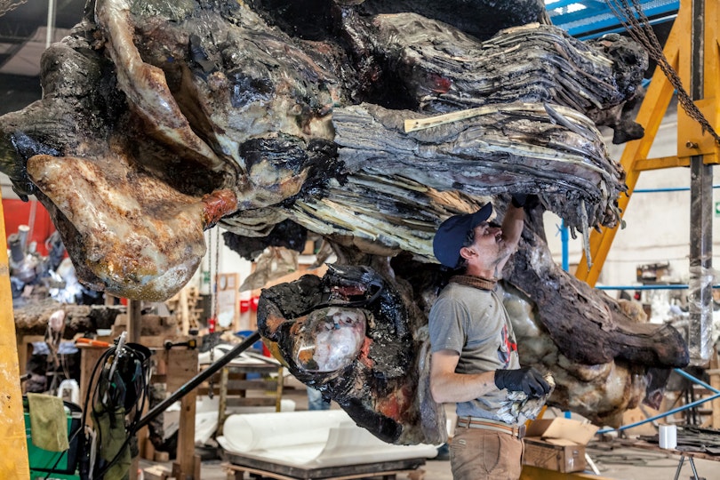 A person stands under a large sculptural form in a workshop space