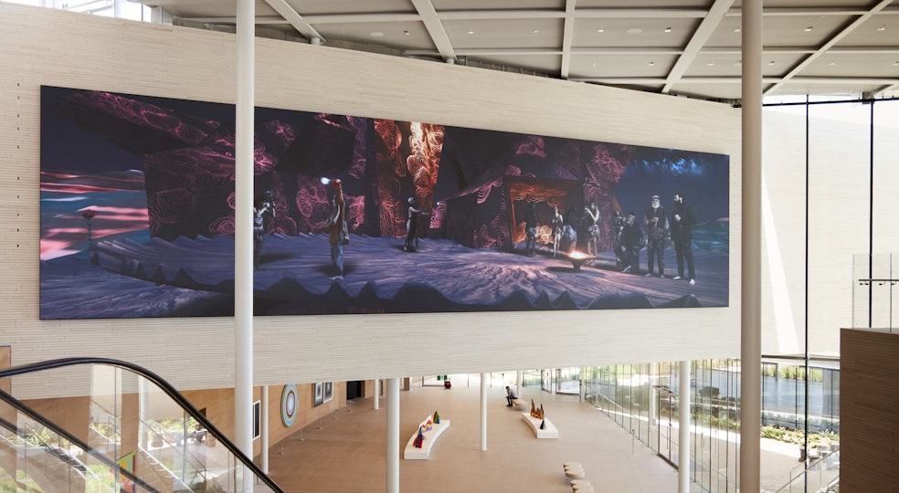An enormous, very wide video screen showing people in an outdoor setting hangs on a wall above an atrium in which other artworks are displayed